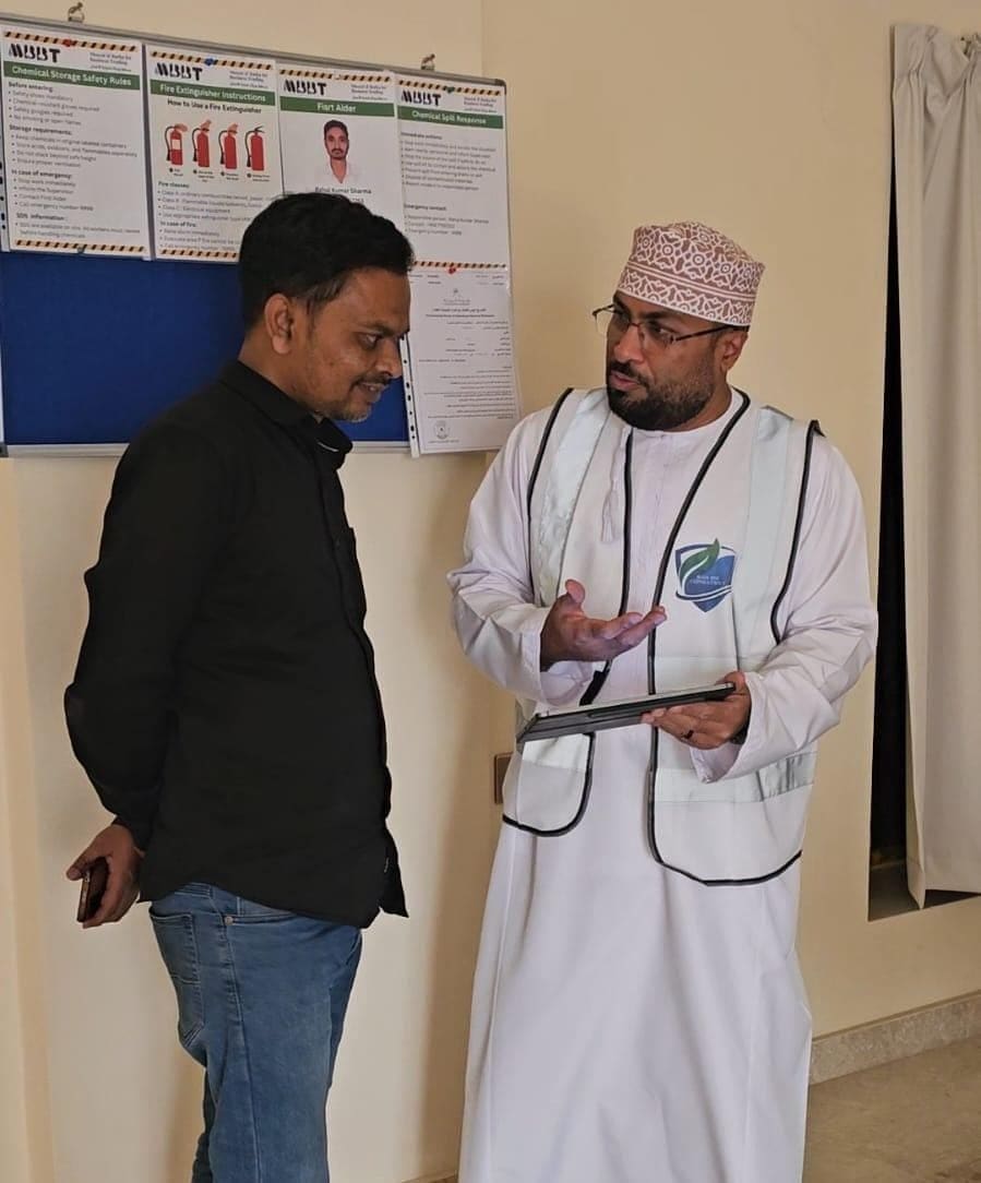Anwar Al-Balushi, in a KADI high-visibility vest, briefing an MBBT team member from a tablet beside the site's safety notice board.